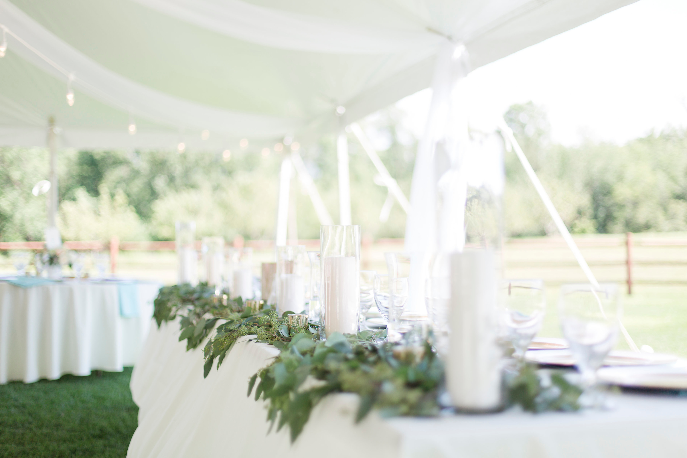 A wedding table decorated with white table cloths and greenery surrounding white tall clear glass candle holders.