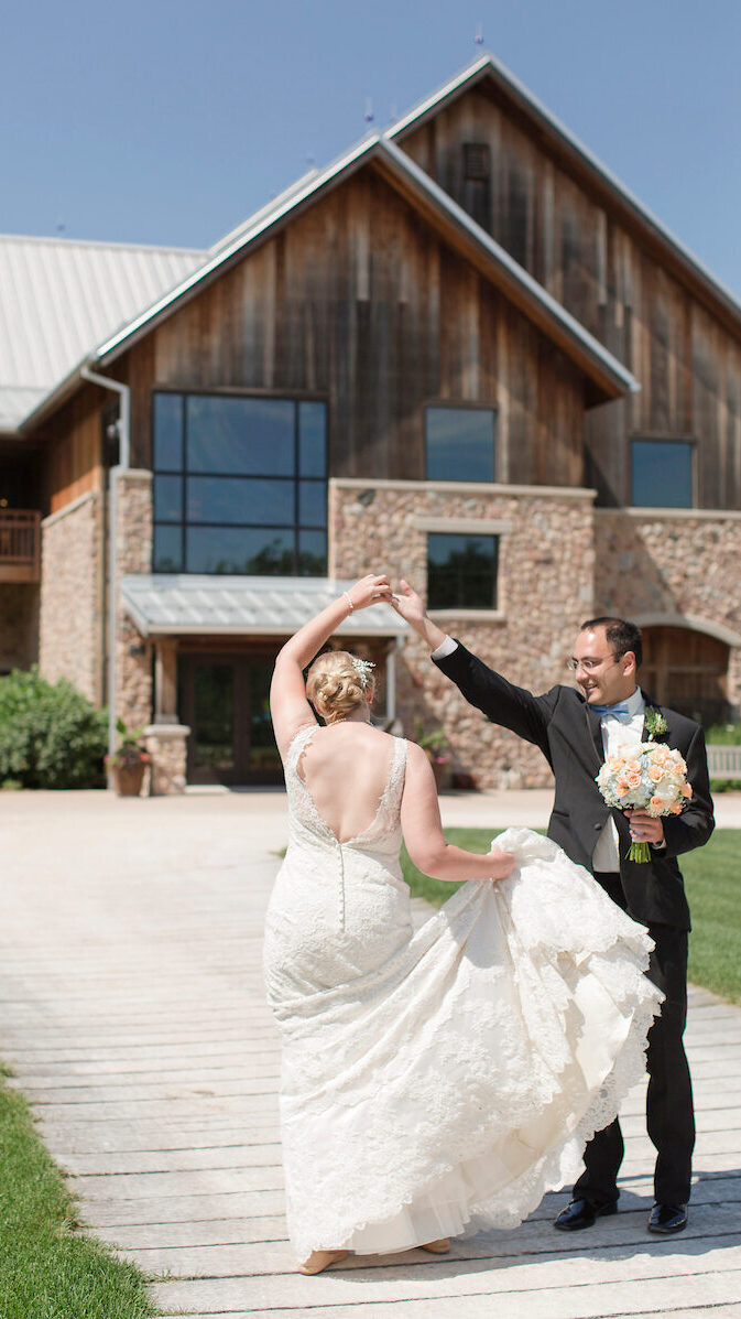 A bride in a floor length backless lace dress is spun around by her groom in a traditional black tux on a cobblestone walkway in front of a the building before their wedding.