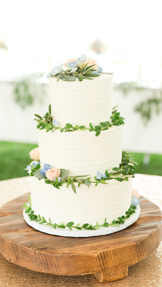 A beautiful 3 tier wedding cake with greenery piped on to each tier's top and mostly white frosting covering the whole cake. There are a few blue and pink roses on the first tier and bottom tier.