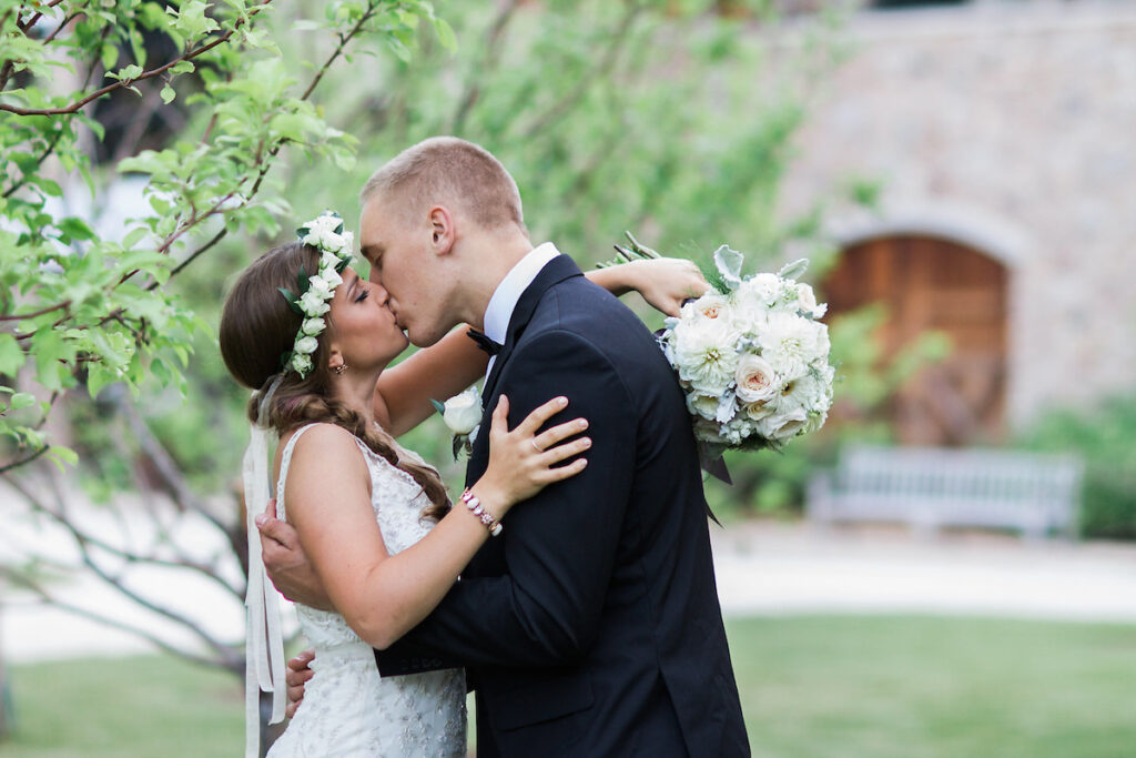A bride and groom, both white, embrace and kiss in front of some trees and a building is in the background.