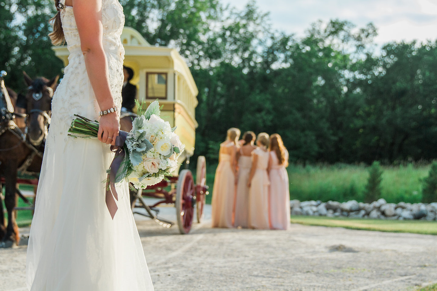 A bride stands in the foreground wearing a white lace dress holding her bouquet. Her bridesmaids stand in the background in their pale pink/peach dresses next to the beige stage coach looking like they are waiting for the bouquet to be thrown behind them. 