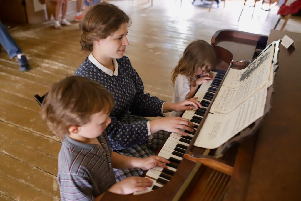 A woman helps two children play piano during her employment
