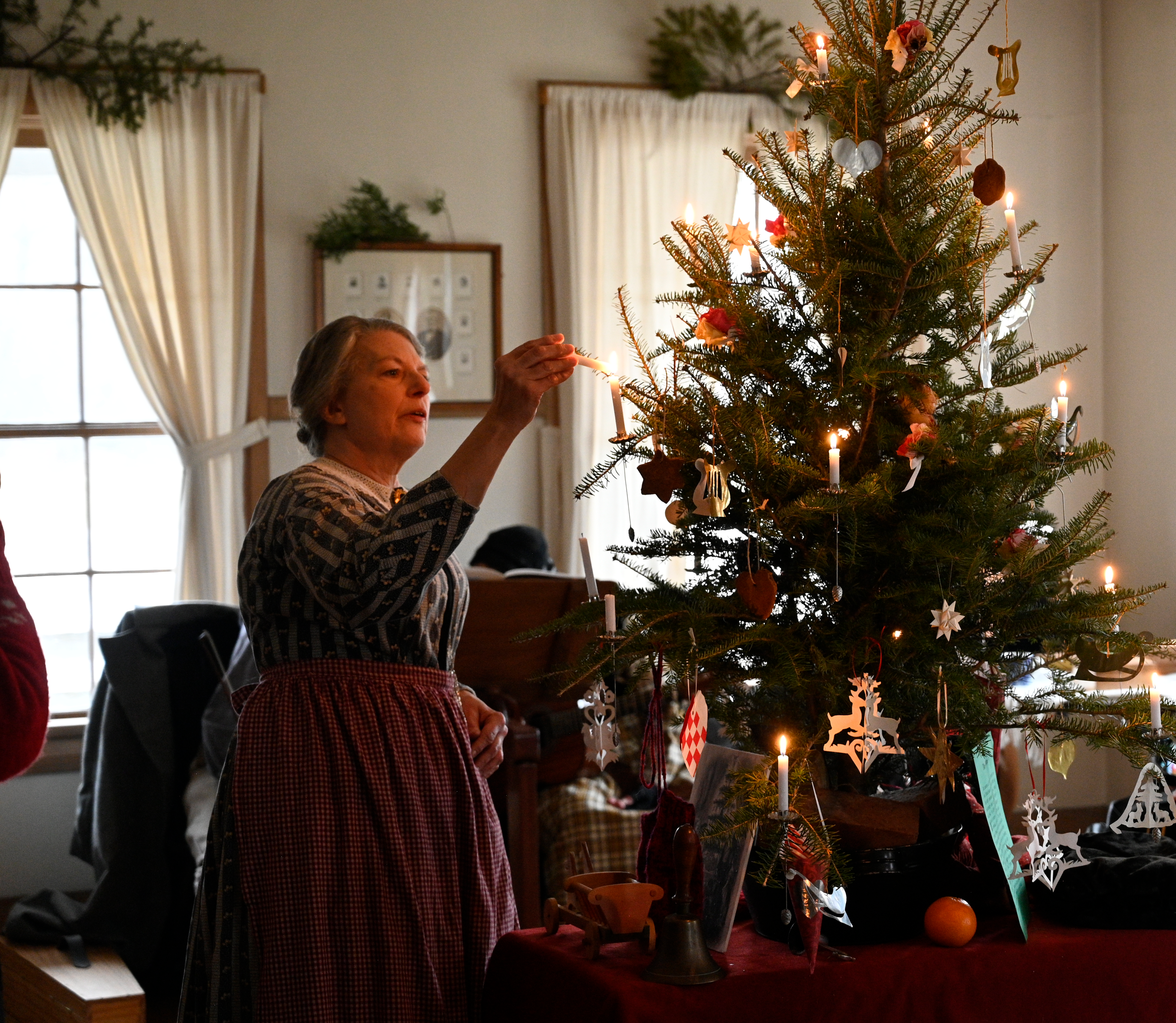 A fair complexioned woman, dressed in period clothing, lighting a small candle on a decorated Christmas tree.